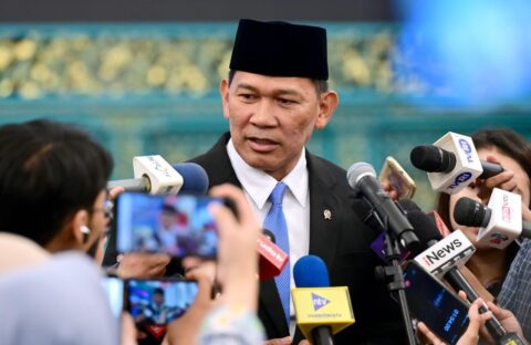 An Indonesian official in a black cap answers questions from a crowd of television reporters holding microphones and phones at the State Palace in Jakarta