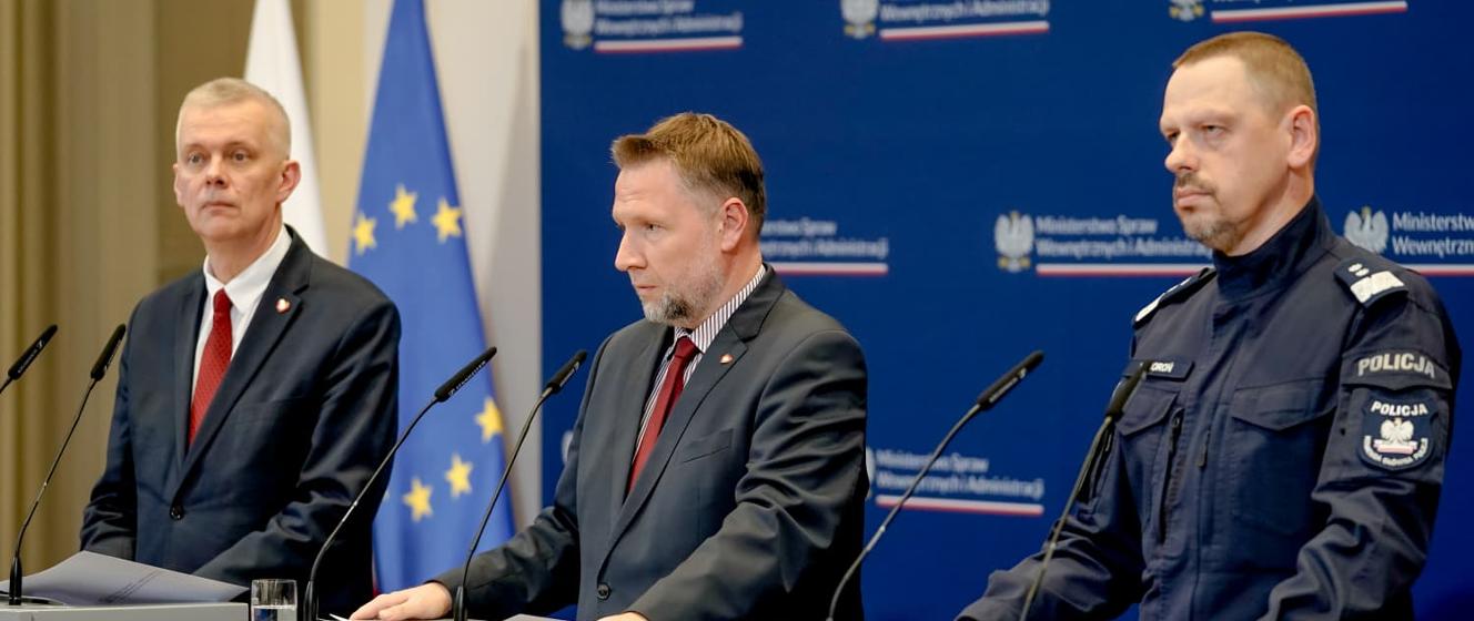 Three officials at microphones in front of Polish Interior Ministry banners, one in police uniform