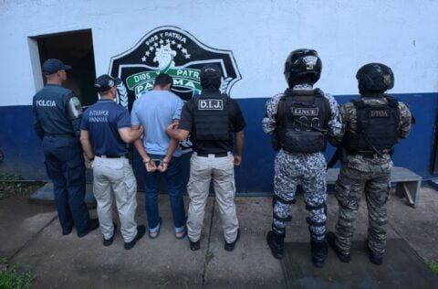 Panamanian police and INTERPOL Panama officers stand with a handcuffed suspect facing a wall during an arrest