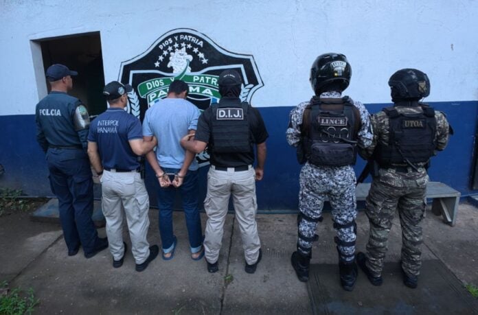 Panamanian police and INTERPOL Panama officers stand with a handcuffed suspect facing a wall during an arrest