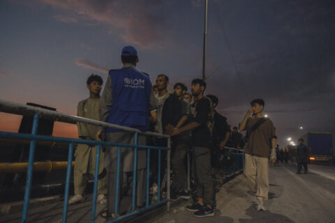 An IOM staff member in a blue vest stands with a group of young men beside a railing at dusk in Afghanistan