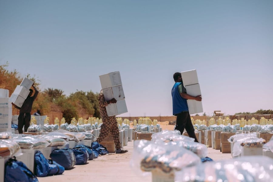 Three workers carry stacked white boxes of aid supplies past rows of bagged relief items under a clear sky