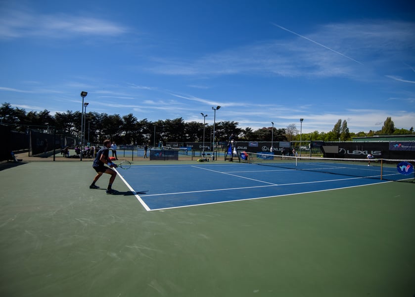 A player waits to receive on a blue hard court with World Tennis Tour banners and an umpire's chair in the background