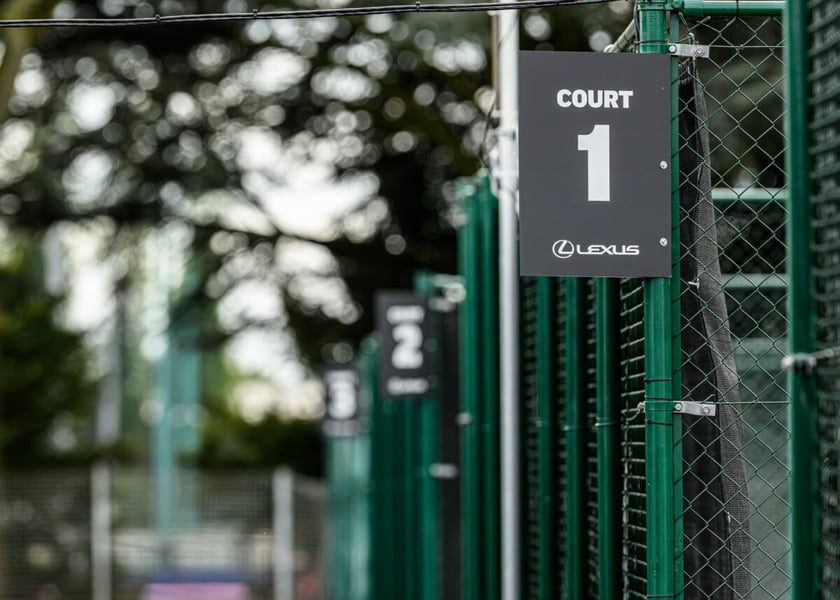 Black Court 1 and Court 2 number signs on a green fence at a tennis venue, with trees blurred behind