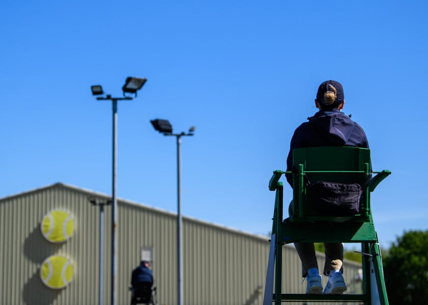 A chair umpire in a navy cap and jacket seen from behind in a green umpire's chair under a blue sky