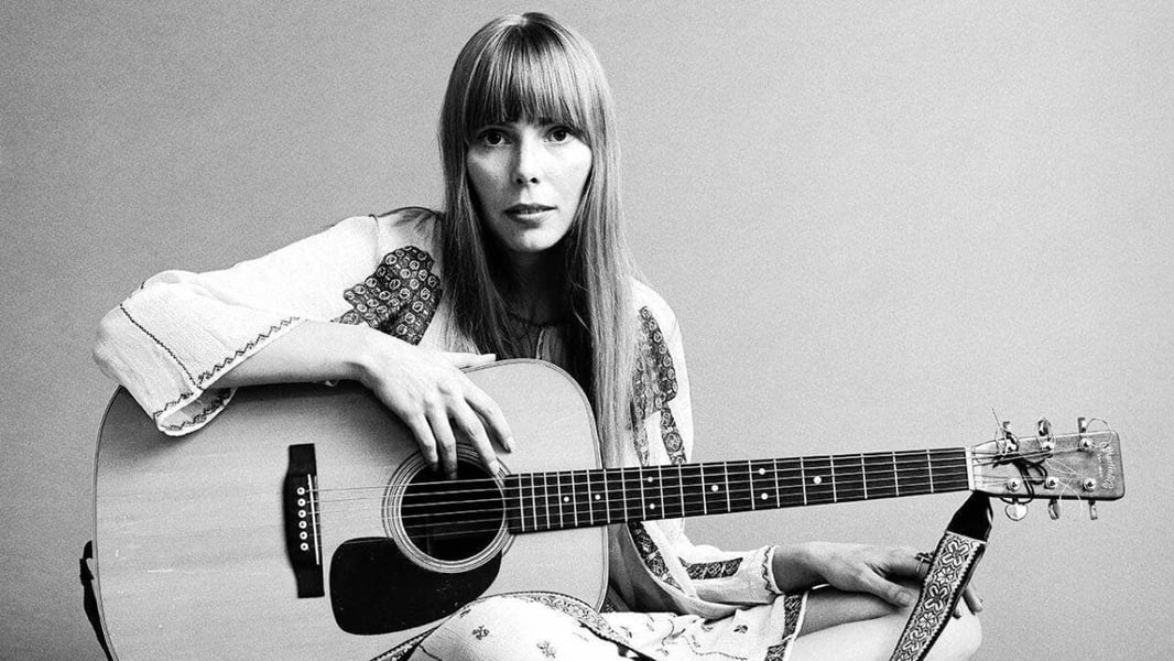 Black-and-white archive portrait of a young Joni Mitchell seated with an acoustic guitar