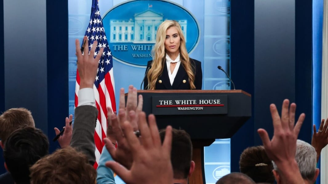 White House press secretary Katie Zacharia at the briefing room podium as reporters raise their hands