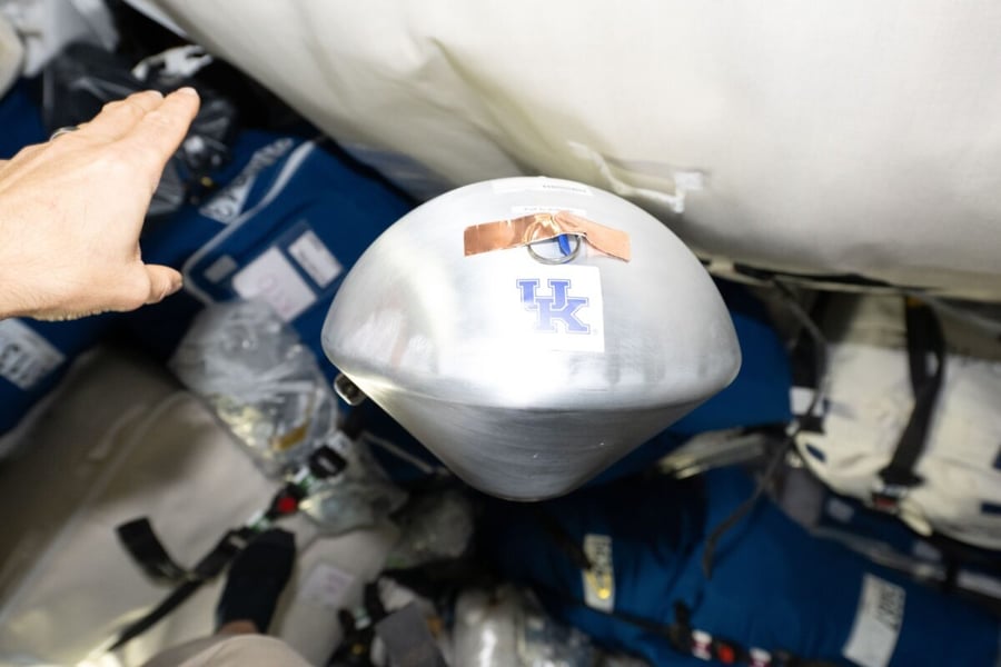 A silver cone-shaped capsule marked with a University of Kentucky logo floats among cargo bags inside the Cygnus XL spacecraft