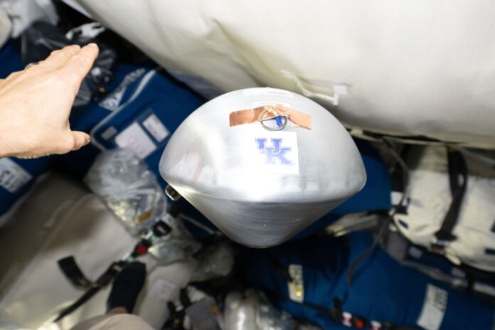A silver cone-shaped capsule marked with a University of Kentucky logo floats among cargo bags inside the Cygnus XL spacecraft
