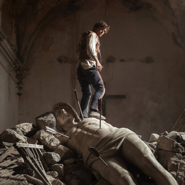 A figure in period clothing stands on a fallen stone statue pierced by arrows amid rubble inside a ruined stone building