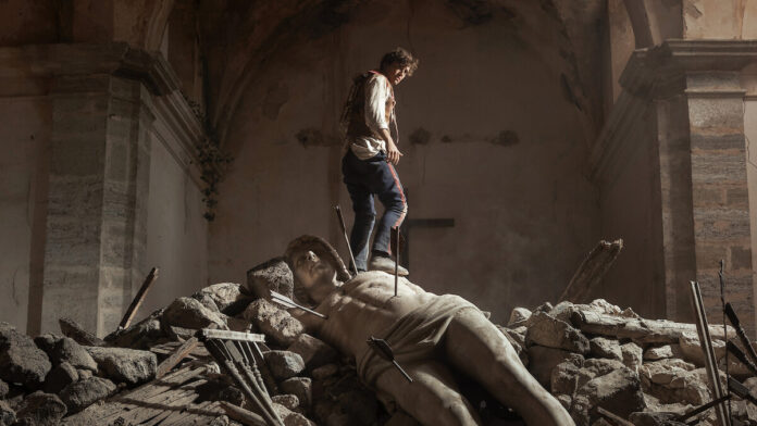 A figure in period clothing stands on a fallen stone statue pierced by arrows amid rubble inside a ruined stone building