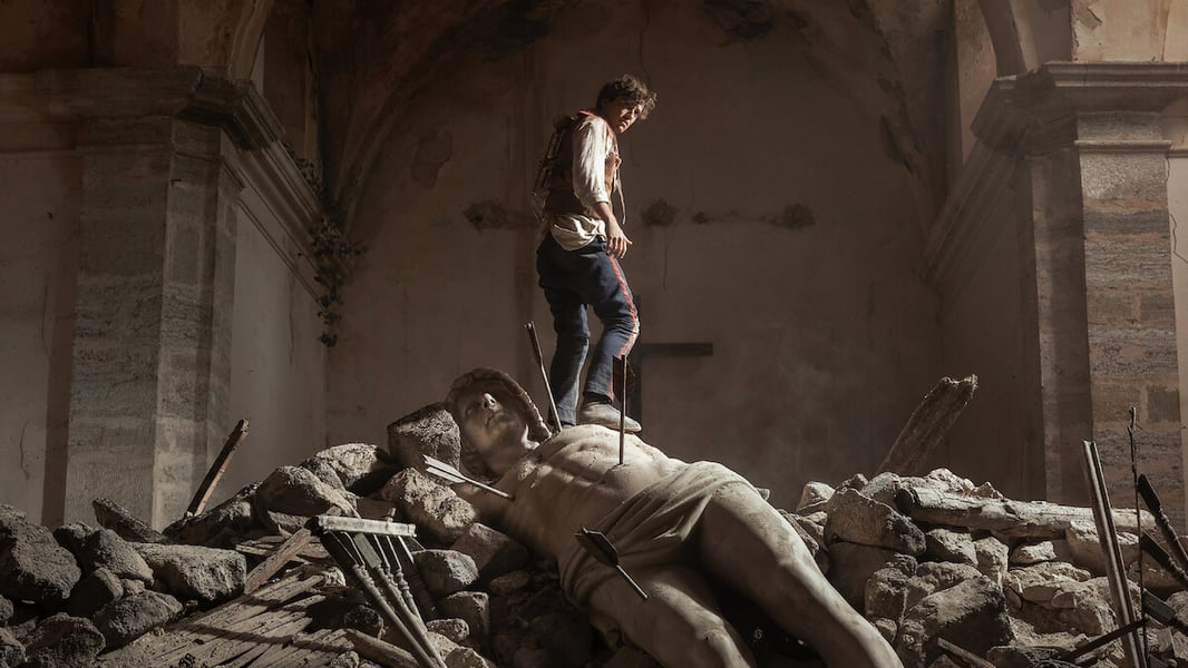 A figure in period clothing stands on a fallen stone statue pierced by arrows amid rubble inside a ruined stone building