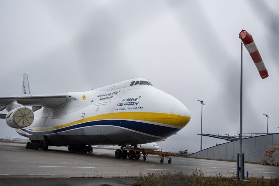 A white Antonov An-124-100M cargo aircraft with blue and yellow livery parked on the apron at Leipzig/Halle Airport beside a windsock
