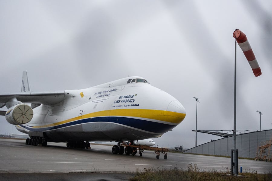 A white Antonov An-124-100M cargo aircraft with blue and yellow livery parked on the apron at Leipzig/Halle Airport beside a windsock