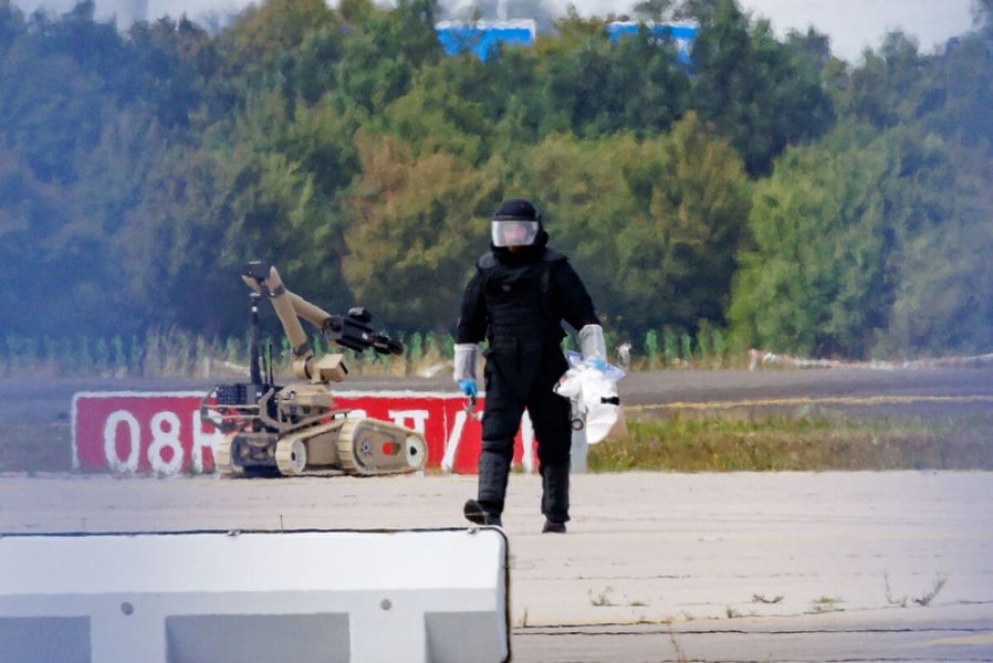 A figure in black protective gear walks across the tarmac at Leipzig/Halle Airport with a bomb-disposal robot visible behind, in front of a tree line