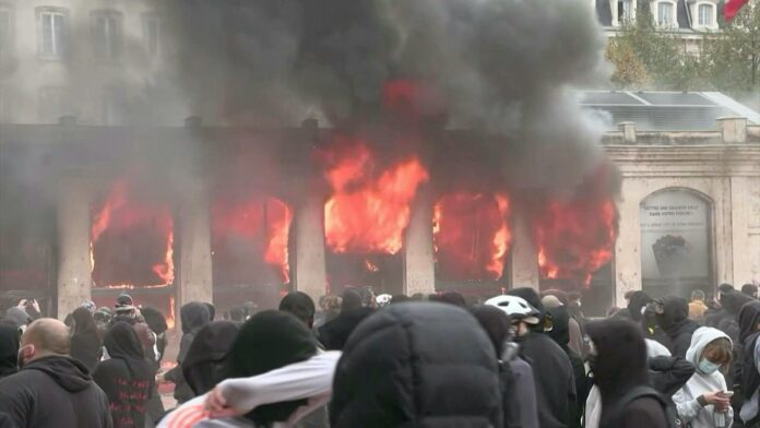 Flames and black smoke pour from the windows of the Lyon tourist office as hooded protesters gather in the square in front