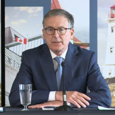 Bank of Canada Governor Tiff Macklem seated at a table taking questions from reporters in Halifax, with lighthouse banners behind him