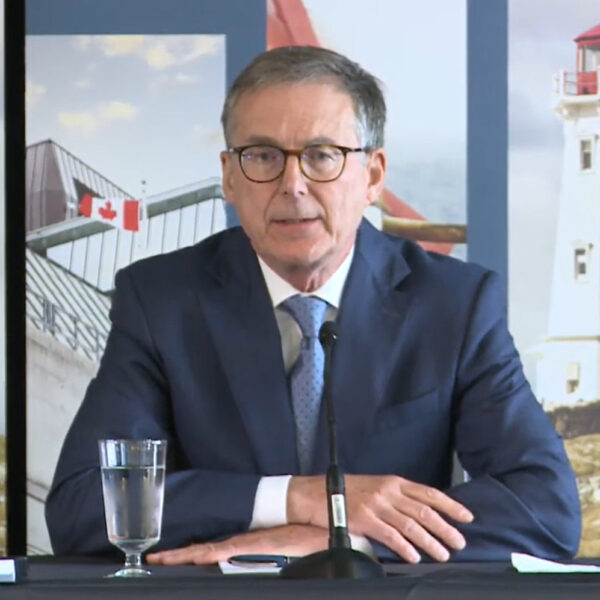 Bank of Canada Governor Tiff Macklem seated at a table taking questions from reporters in Halifax, with lighthouse banners behind him