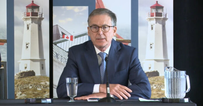 Bank of Canada Governor Tiff Macklem seated at a table taking questions from reporters in Halifax, with lighthouse banners behind him