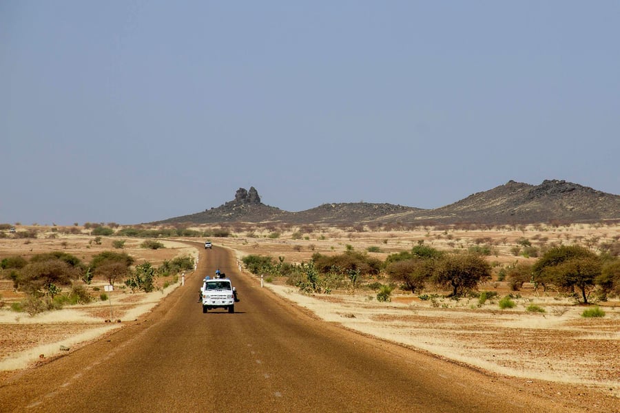 A white UN vehicle travels a paved desert road toward rocky hills in northern Mali