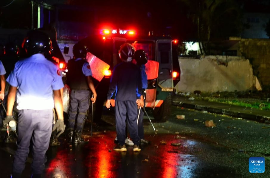 Police officers in helmets and shields beside a vehicle with flashing lights on a dark street strewn with debris
