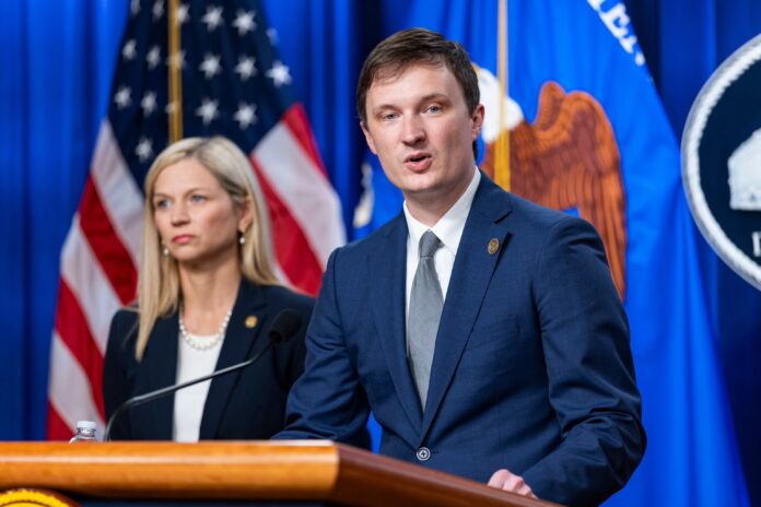 Acting Deputy Attorney General Trent McCotter speaks at a lectern at the Justice Department, with a US flag behind him
