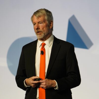 Strategy Executive Chairman Michael Saylor in a dark suit and orange tie speaking on stage
