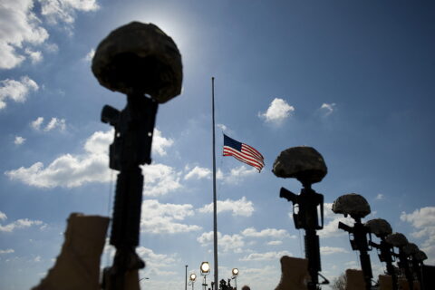 Memorial rifles topped with combat helmets in front of a US flag flying at half-staff