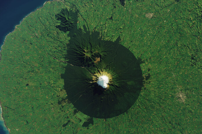 Landsat 8 satellite image of snow-capped Mount Taranaki in New Zealand ringed by dark forest inside a circular protected-area boundary, surrounded by green farmland