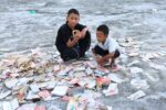 Two boys crouch among scattered school supplies on grey flood sediment in Nuwakot, Nepal