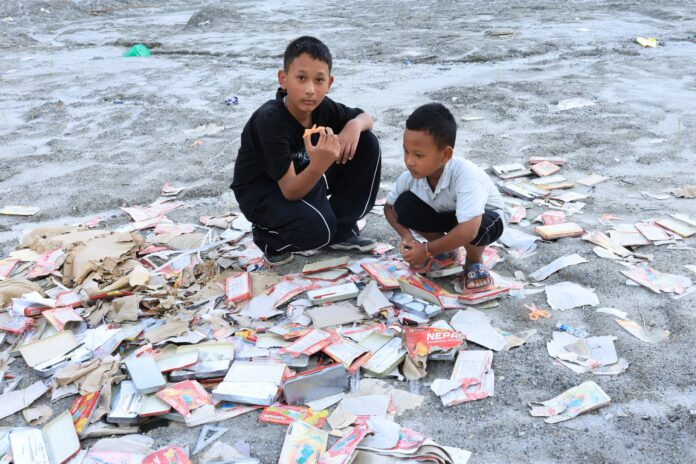 Two boys crouch among scattered school supplies on grey flood sediment in Nuwakot, Nepal