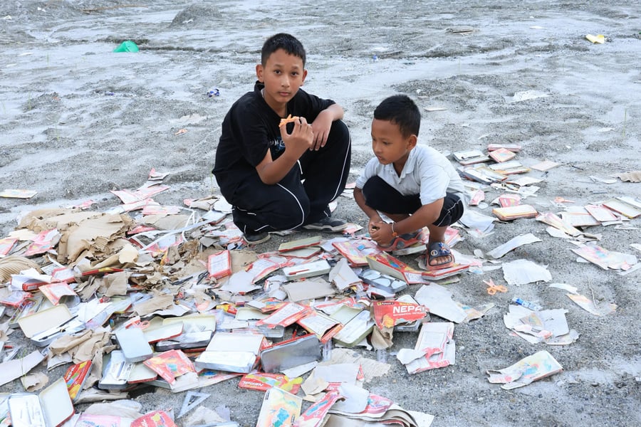 Two boys crouch among scattered school supplies on grey flood sediment in Nuwakot, Nepal