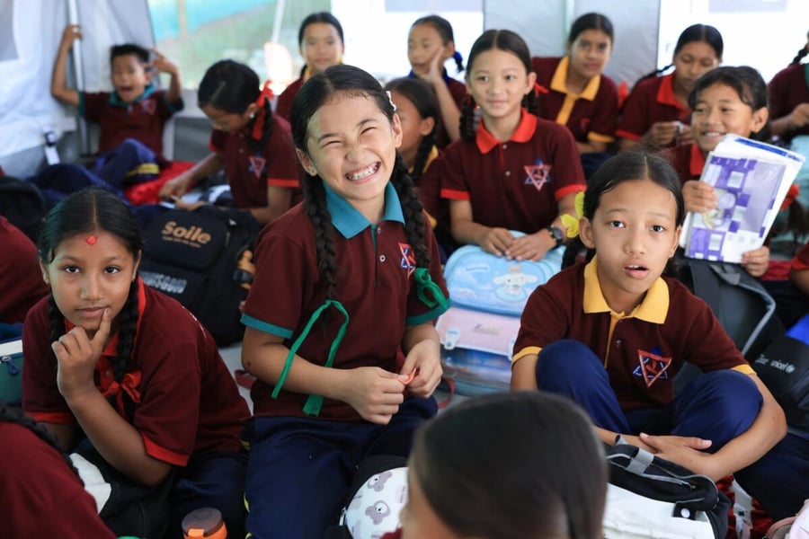 Girls in maroon school uniforms sit close together in a temporary tent classroom in Nepal