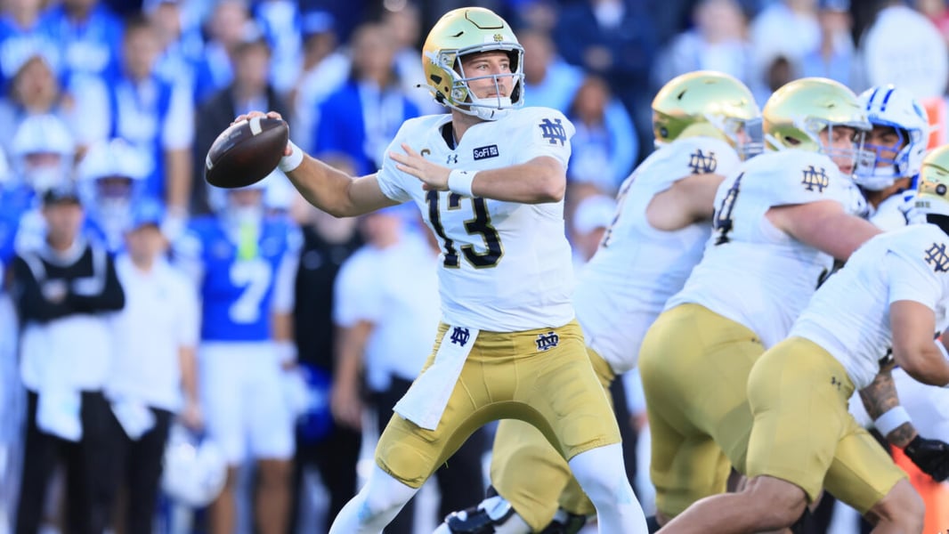 A Notre Dame quarterback in a white No. 13 jersey cocks his arm to throw as linemen block and BYU fans watch from behind