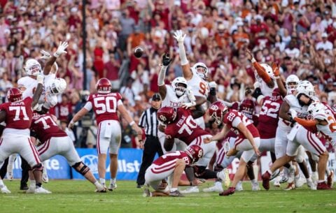 An Oklahoma kicker in a white uniform kicks a field goal as two Texas defenders in white and red leap to try to block it during the Red River Rivalry