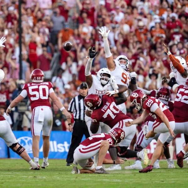 An Oklahoma kicker in a white uniform kicks a field goal as two Texas defenders in white and red leap to try to block it during the Red River Rivalry