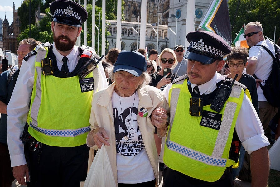 Two Metropolitan Police officers lead an elderly woman away at a Palestine Action protest in Parliament Square