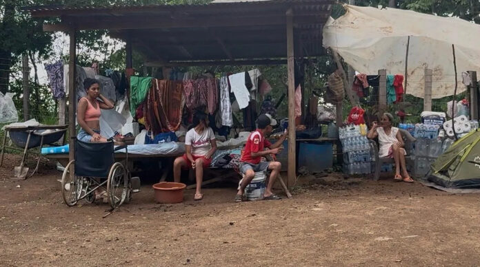 Families sitting under a makeshift shelter with laundry hanging and water bottles stacked beside them