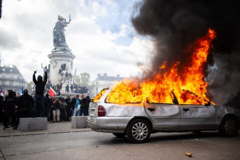 A silver estate car burns with tall flames and black smoke while masked protesters stand near a monument in Paris