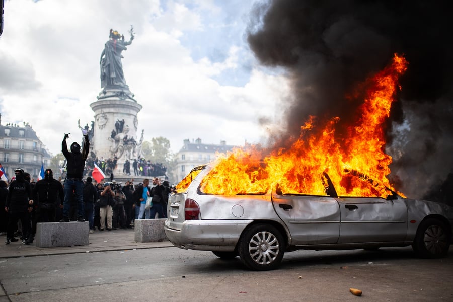 A silver estate car burns with tall flames and black smoke while masked protesters stand near a monument in Paris