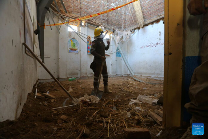 A rescuer in a yellow helmet stands with a shovel in a classroom with the roof torn open after a school roof collapse in Peshawar