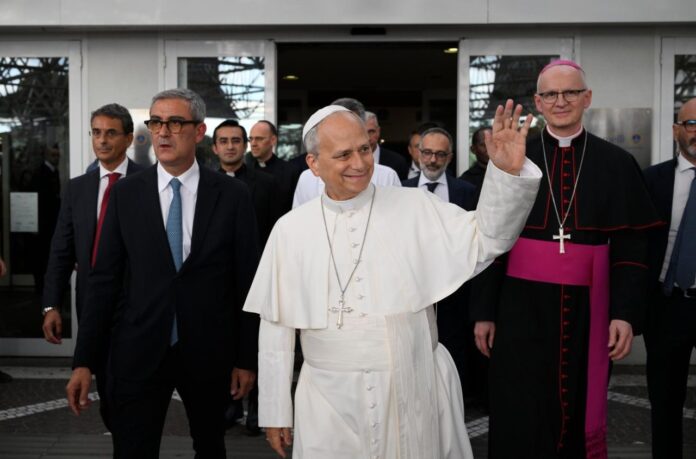 Pope Leo XIV in white waves with his right hand as he walks out of Gemelli Hospital, flanked by men in suits and a bishop