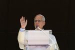 Pope Leo XIV raises his right hand in blessing at a clear lectern during the Sunday Angelus