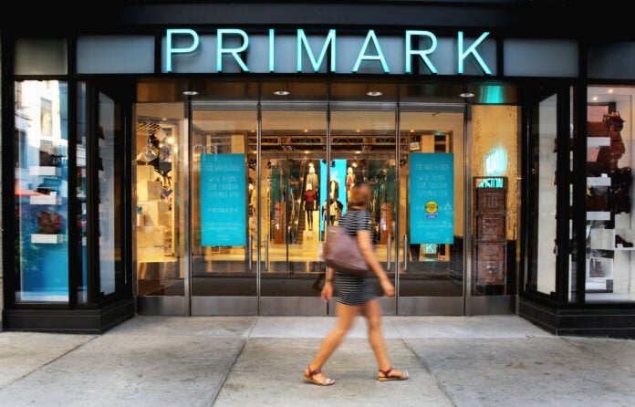 A woman walks past the glass entrance of a Primark store in Boston under the illuminated Primark sign, with posters announcing the opening on September 10
