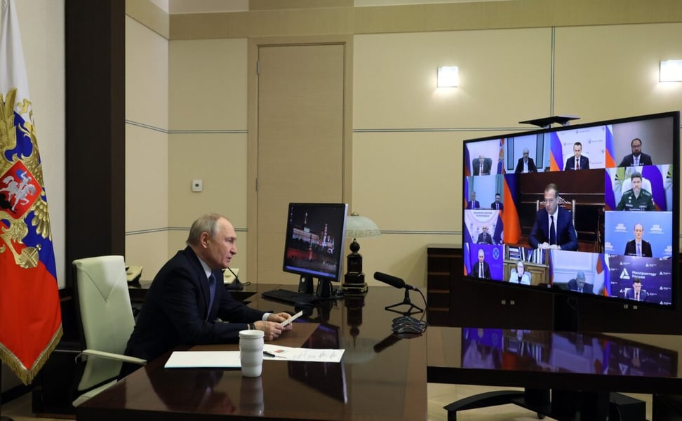 Vladimir Putin at his desk with participants of the information security meeting shown on a screen beside him