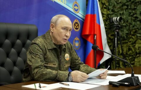 Vladimir Putin in military-style jacket speaks at a table with a microphone and Russian flag behind him at a Joint Force command post