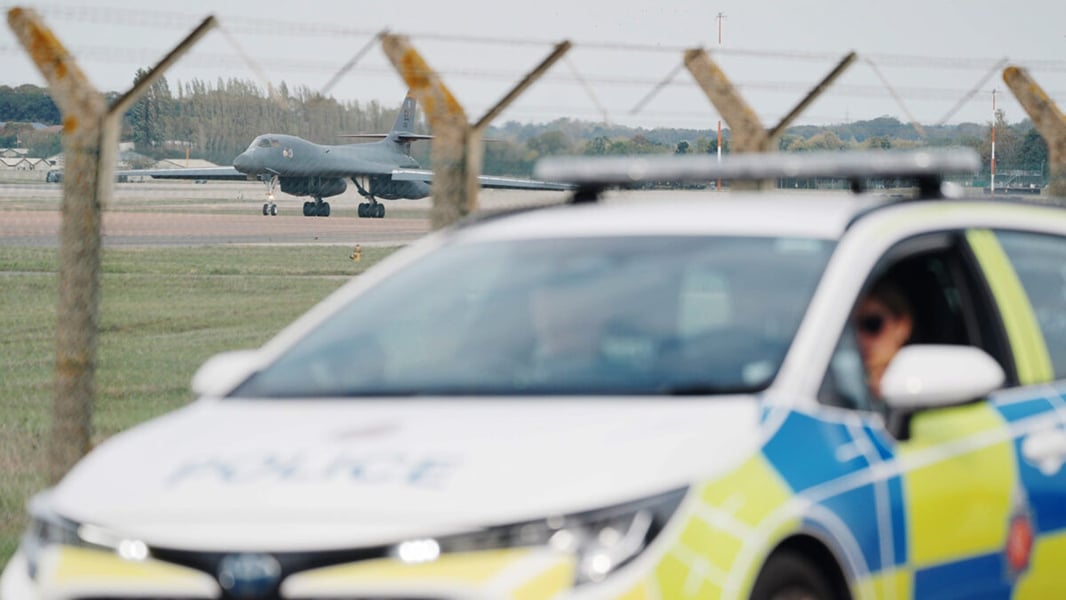 A police car passes the perimeter fence at RAF Fairford as a US Air Force B-1 bomber taxis on the runway behind it