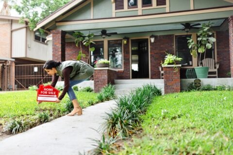 A real estate agent bends to place a red Home For Sale sign in the lawn in front of a brick craftsman-style house
