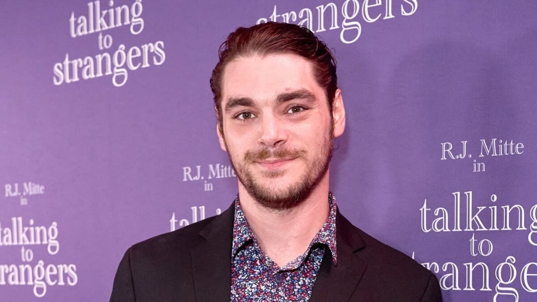 RJ Mitte smiling in front of a purple Talking To Strangers backdrop, wearing a dark jacket and a floral shirt