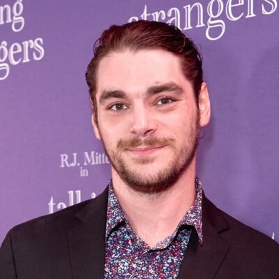 RJ Mitte smiling in front of a purple Talking To Strangers backdrop, wearing a dark jacket and a floral shirt